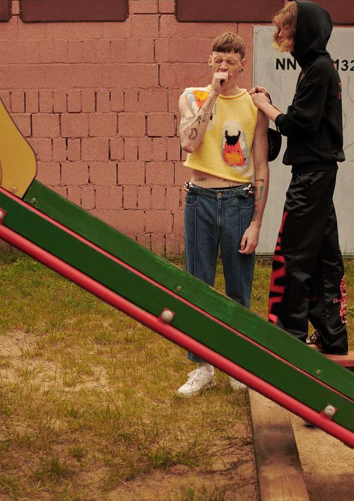 Two young adult males in casual fashion on a playground near a brick wall and slide, captured in natural light.