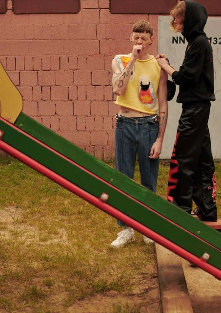 Two young adult males in casual fashion on a playground near a brick wall and slide, captured in natural light. 