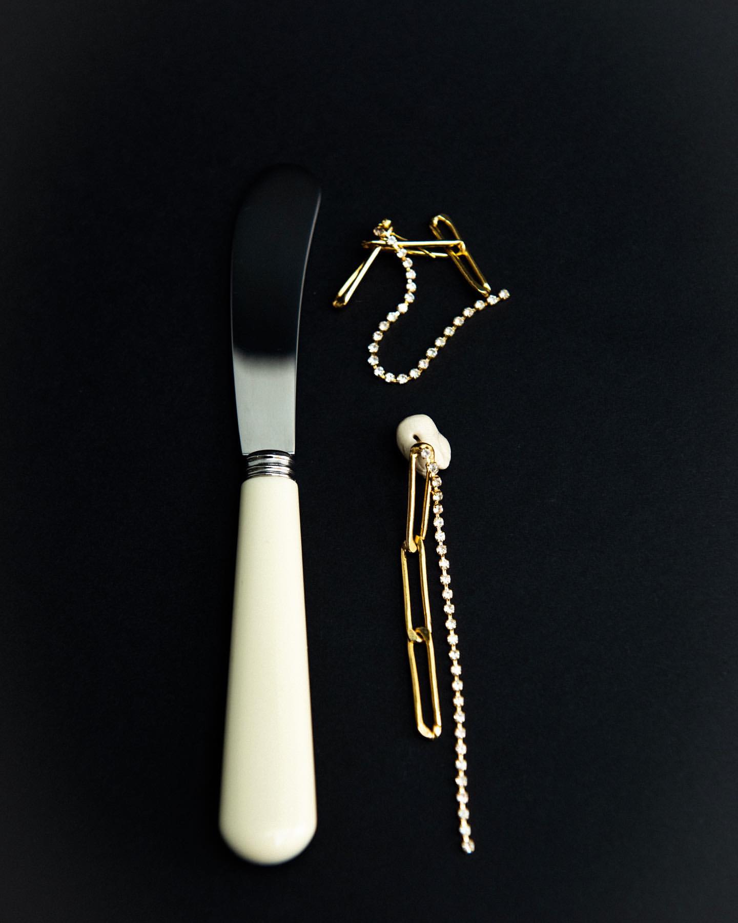 Still life of a silver butter knife with an off-white handle and two gold earrings with crystal accents on a black background.