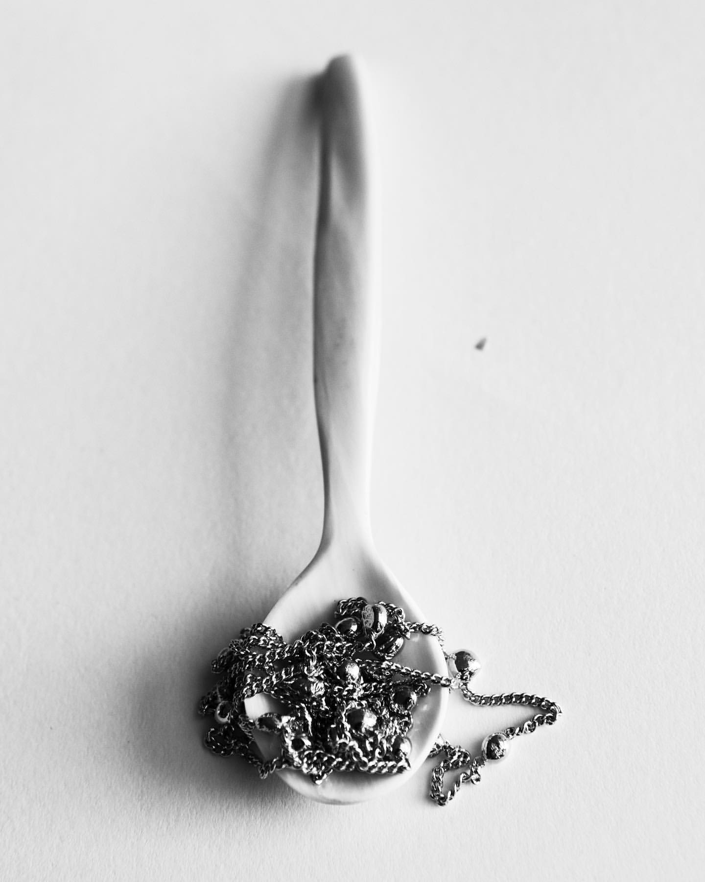 A monochromatic still life presents a spoon filled with a silver chain, all set against a bright white backdrop, minimal composition.