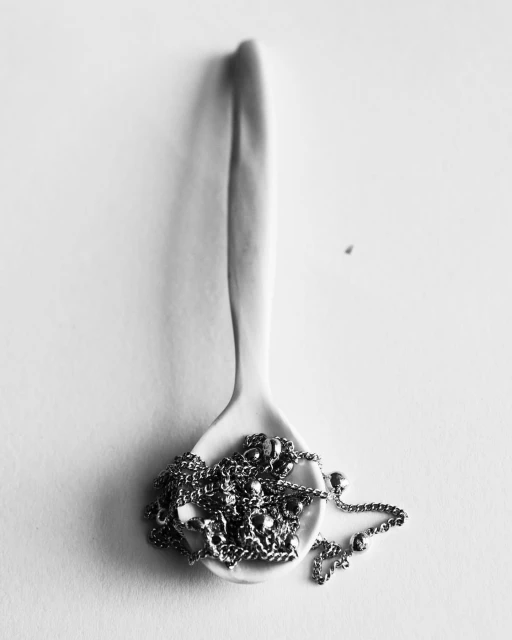 A monochromatic still life presents a spoon filled with a silver chain, all set against a bright white backdrop, minimal composition.