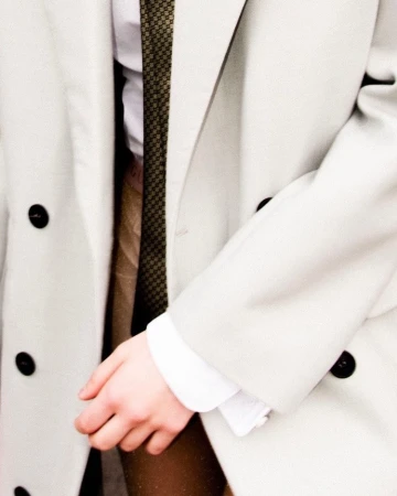 Close-up of a person wearing a white double-breasted coat over a white shirt and patterned tie with brown trousers.