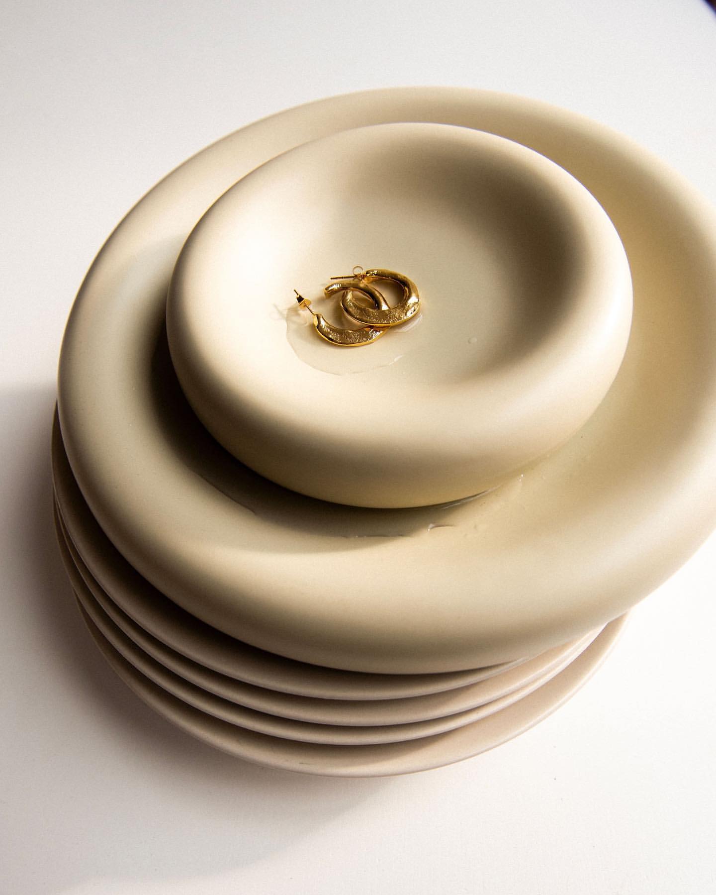 Elegant gold hoop earrings nestled in a ceramic dish, complemented by a stack of matching plates in a minimalist setting.