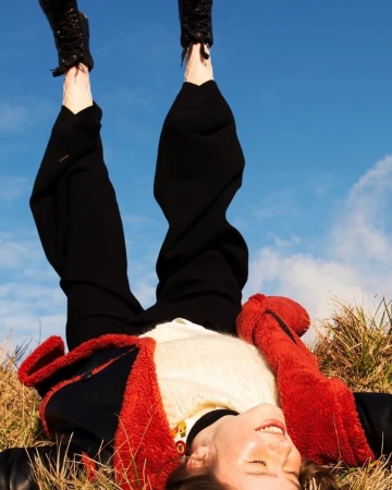 Person lying upside down on dry grass with legs raised against a bright blue sky, wearing black trousers and red jacket.