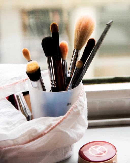 Makeup brushes arranged in a container on a windowsill, casting soft shadows in diffused natural light. Makeup compact visible in foreground.
