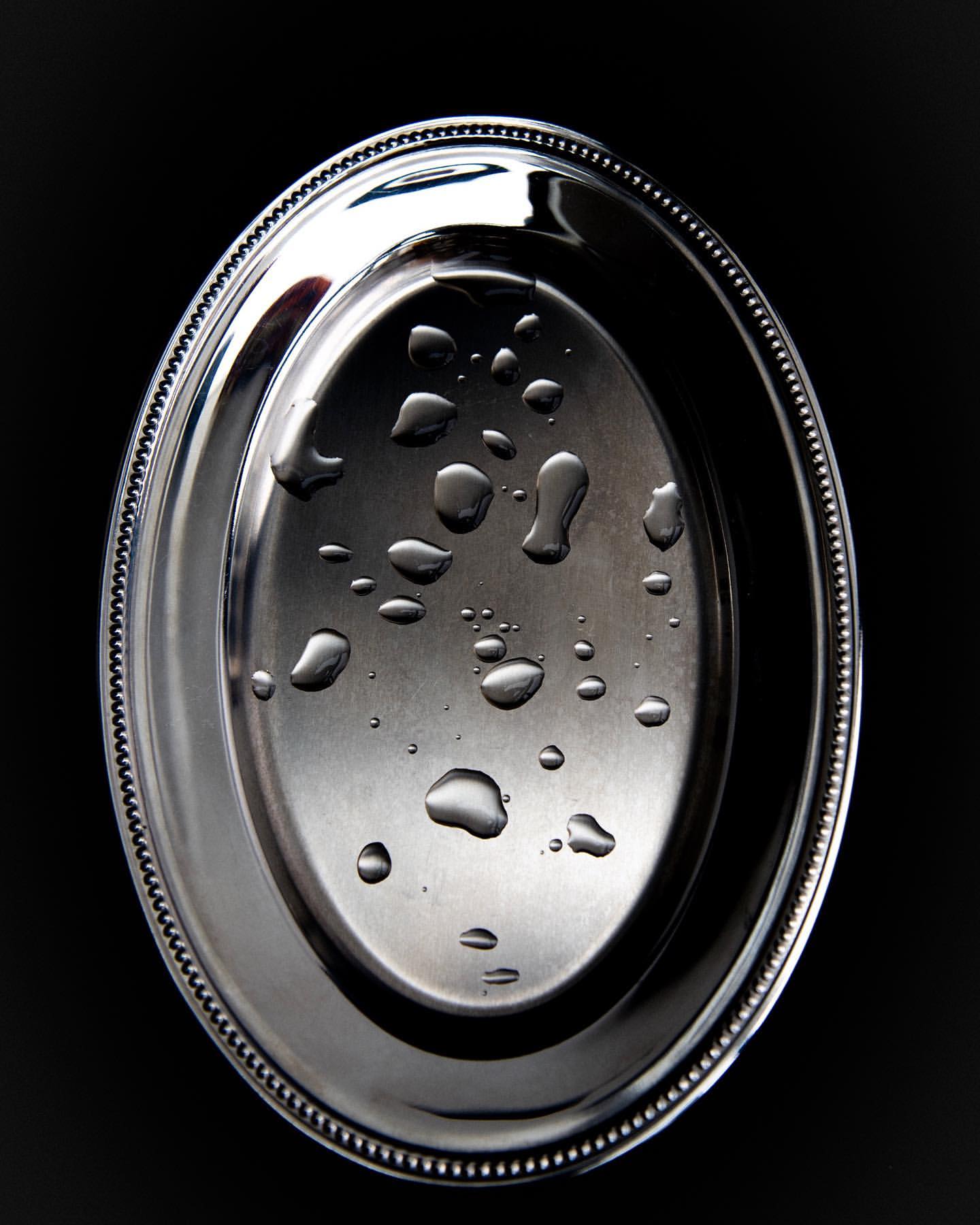 Elevated view of a silver oval serving tray with a beaded rim and water droplets on a black background creating a minimalist food still life.