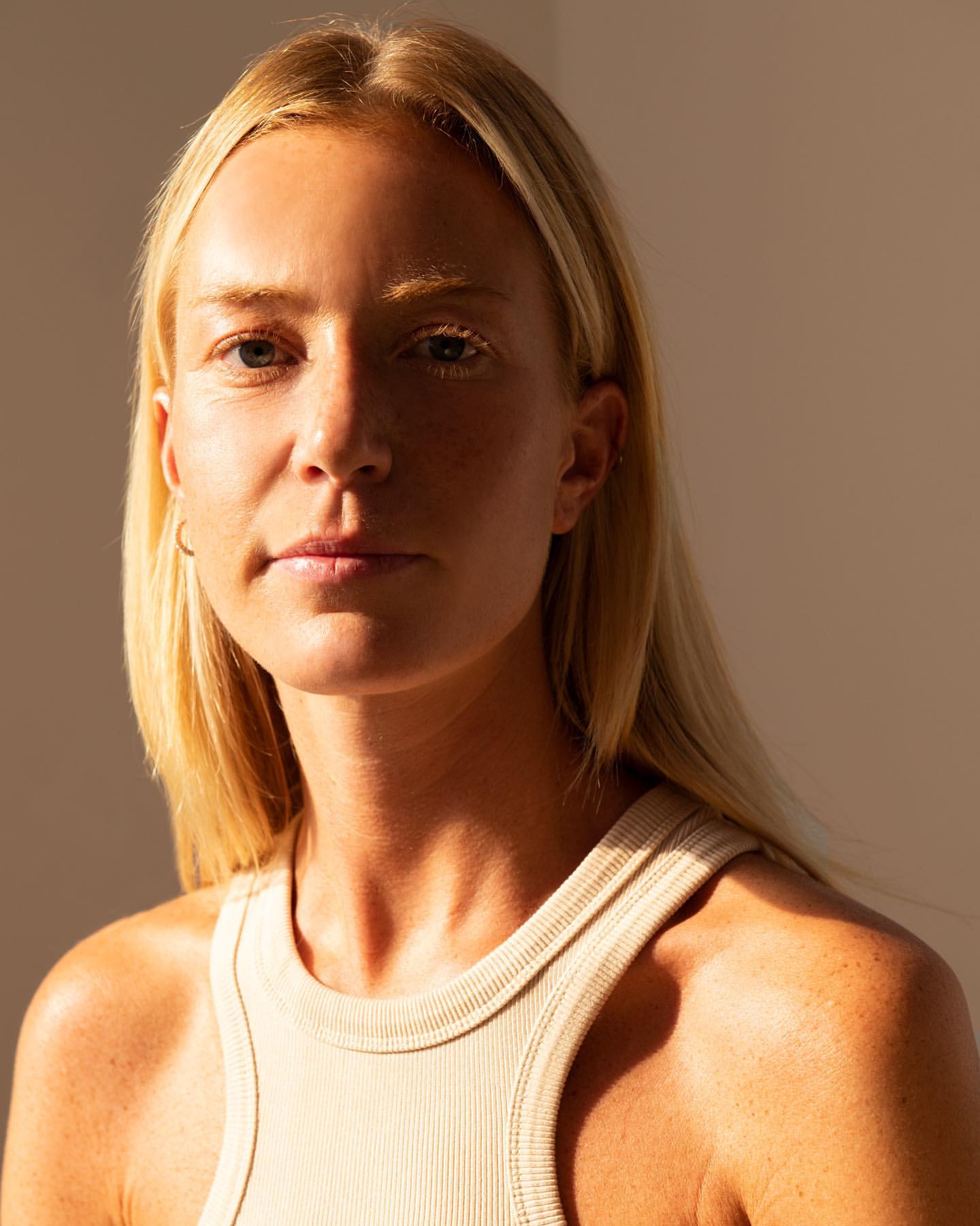 Portrait of a fair-skinned woman with long blonde hair in sunlight, wearing a beige ribbed tank top against a neutral background.