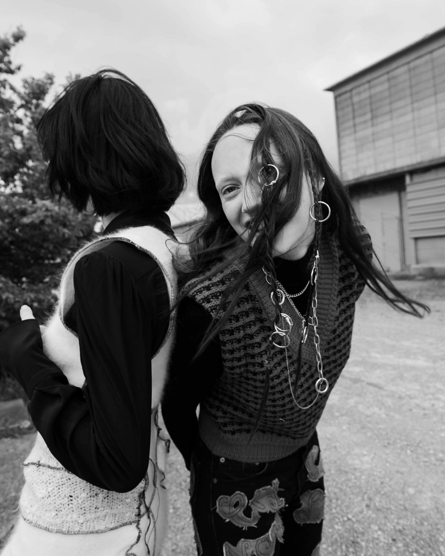 Two young women stand outdoors in vests, accessorized with jewelry. Monochrome shot against a blurred urban background.