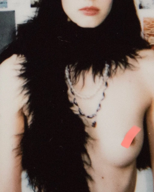 Close-up of a topless adult female wearing a black feather boa and a beaded necklace, indoors with blurred background.