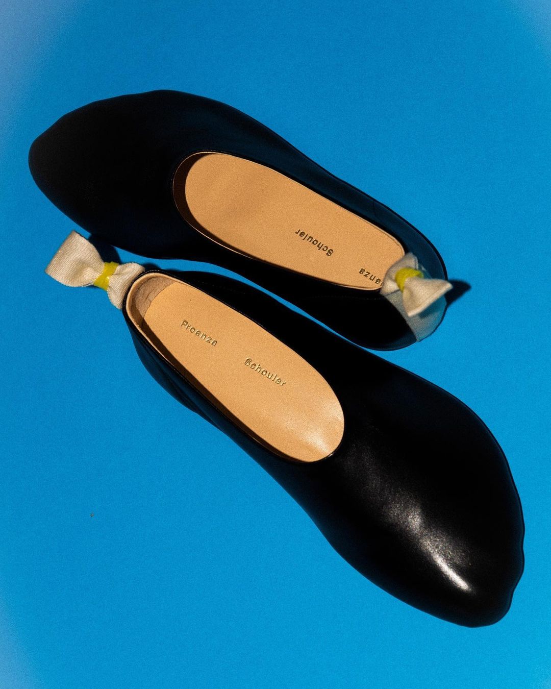 Pair of black leather ballet flats with beige soles and a small bow on a bright blue background for fashion product photography.