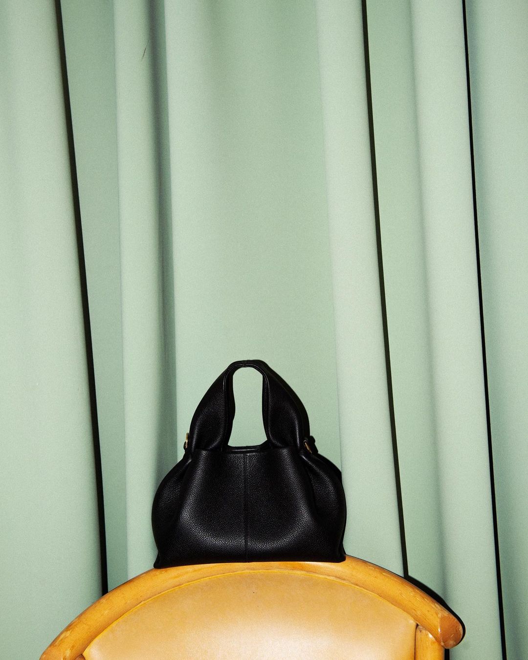 Black leather handbag sits on a gold chair against a mint green curtain backdrop in a simple, minimalist fashion studio photograph.