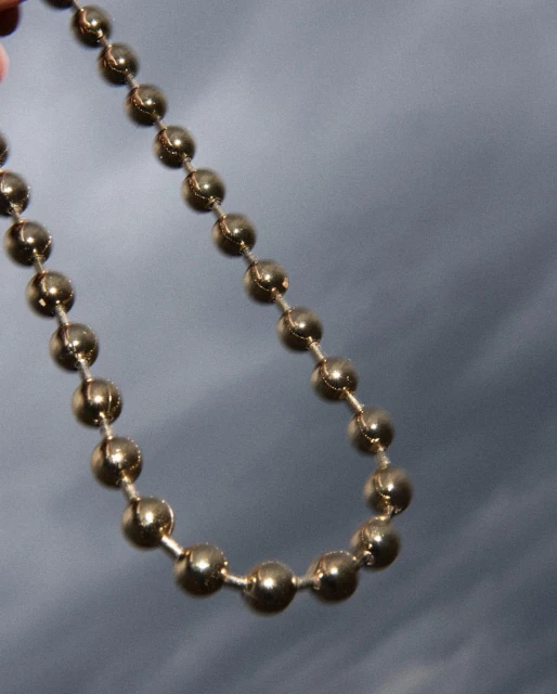 Close-up of a gold-colored beaded chain against a blurred grey background, showcasing its reflective surface and simple design.