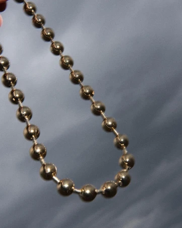 Close-up of a gold-colored beaded chain against a blurred grey background, showcasing its reflective surface and simple design.
