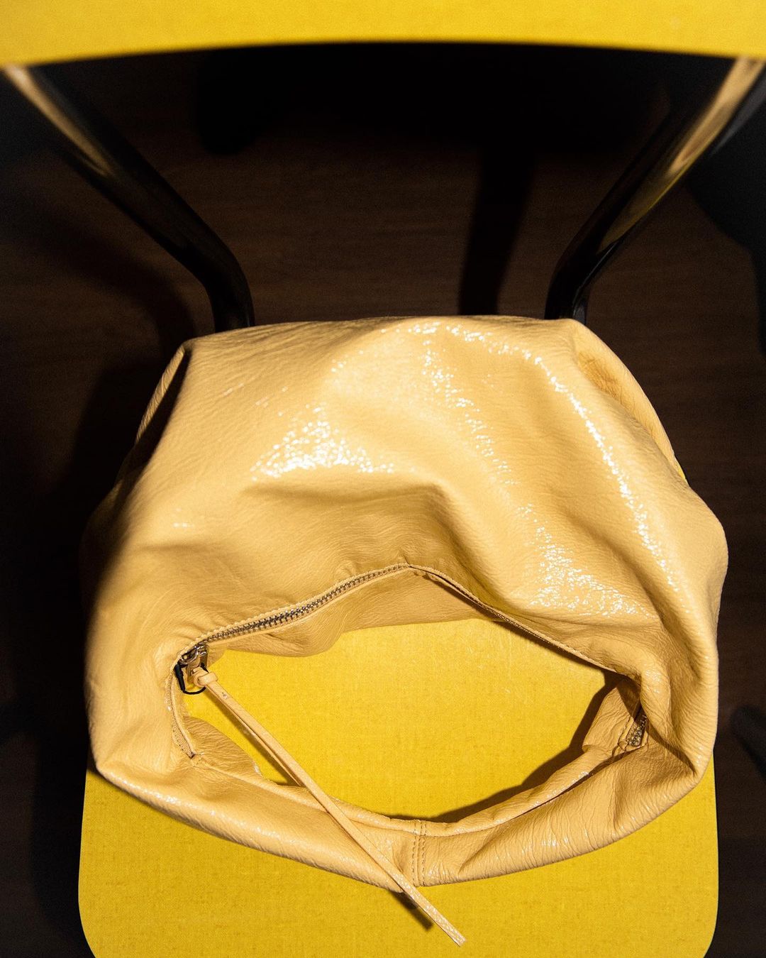 Elevated close-up of a yellow leather shoulder bag resting on a yellow padded chair with a black metal frame, casting deep shadows.