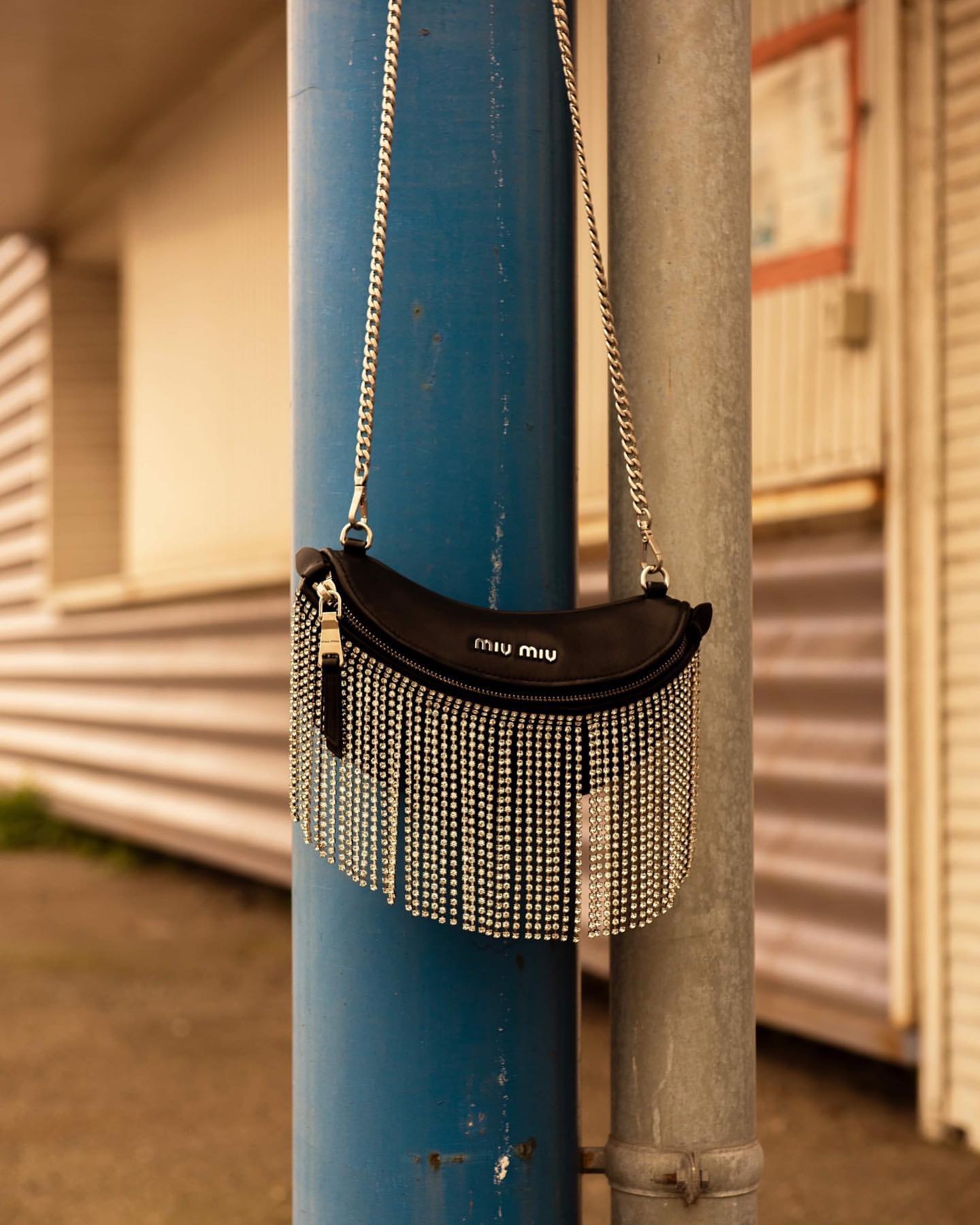 Miu Miu black leather handbag with a silver chain strap and silver fringe detail, showcased against a blue and beige outdoor setting.
