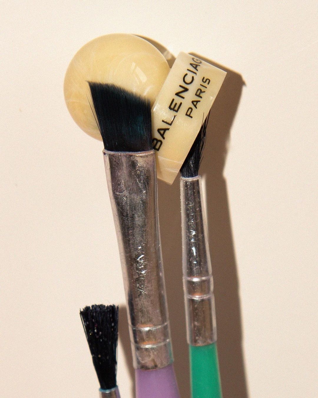 Still life featuring an orb, Balenciaga Paris tag, and an arrangement of small paintbrushes against a neutral backdrop.
