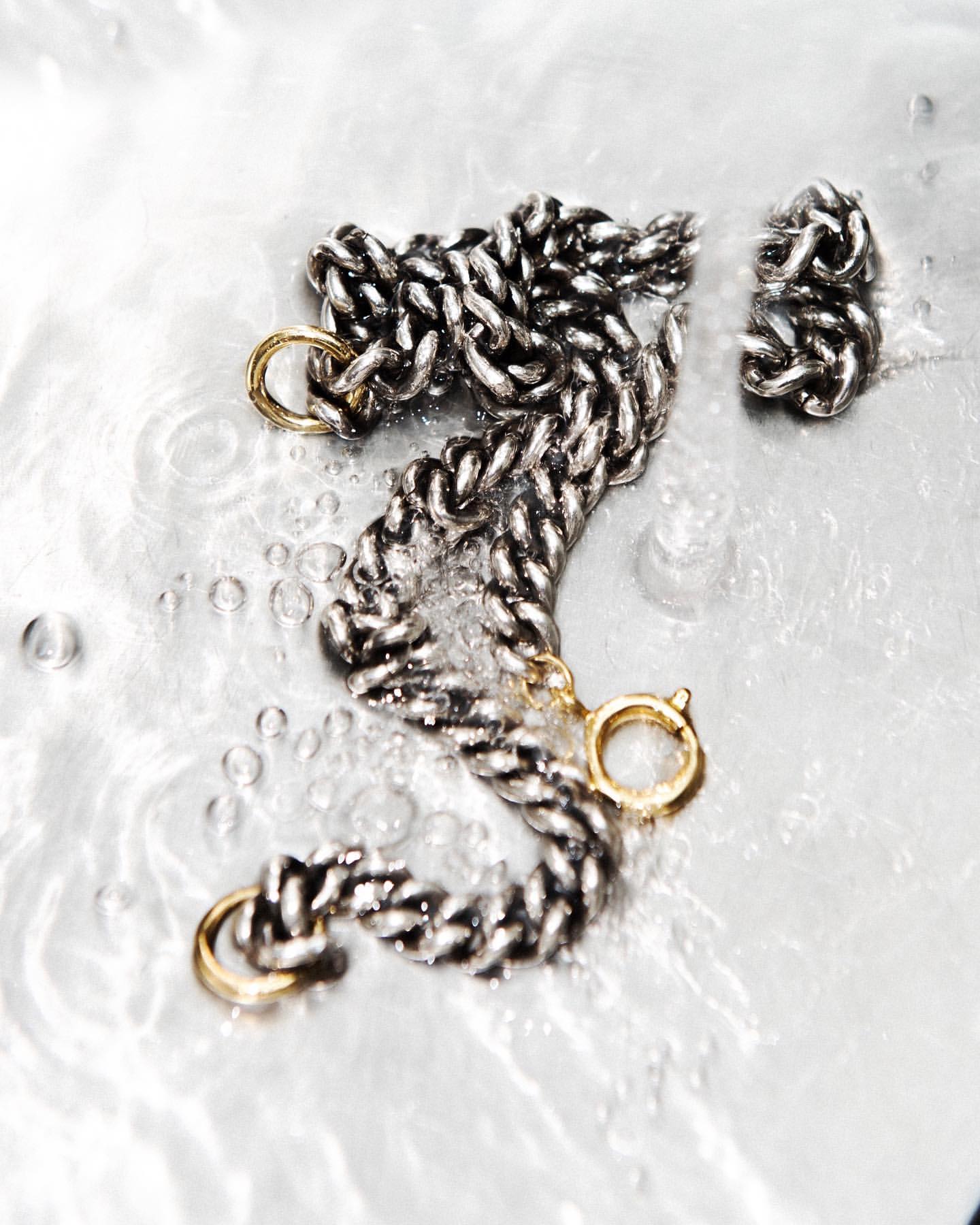 Close-up of a silver chain necklace with gold clasp accents on a reflective surface with water droplets, jewellery, beauty and fashion.