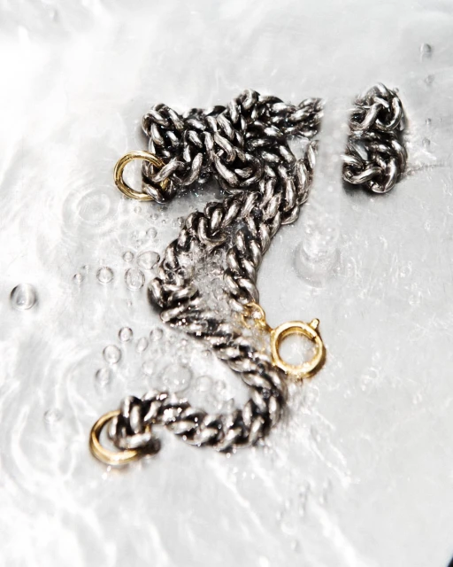 Close-up of a silver chain necklace with gold clasp accents on a reflective surface with water droplets, jewellery, beauty and fashion.