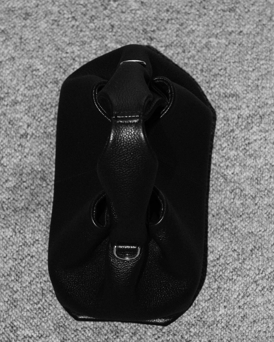 Black leather handbag with a unique handle, displayed on a textured grey background with subtle lighting for a studio-quality look.