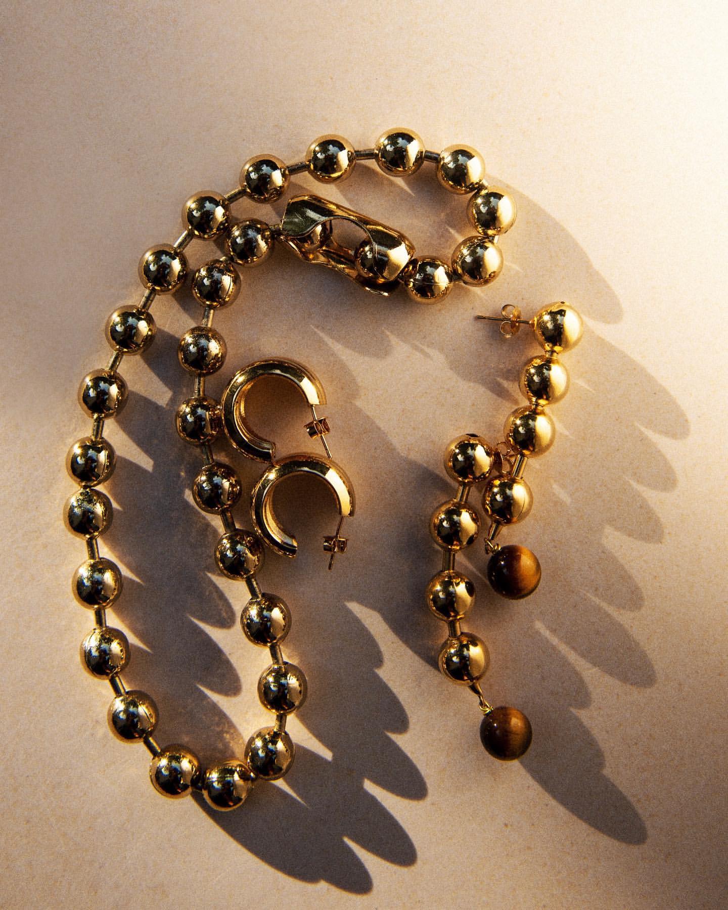 Elegant gold jewelry set: a ball chain necklace, hoop earrings, and drop earrings with tiger eye beads against a beige background.