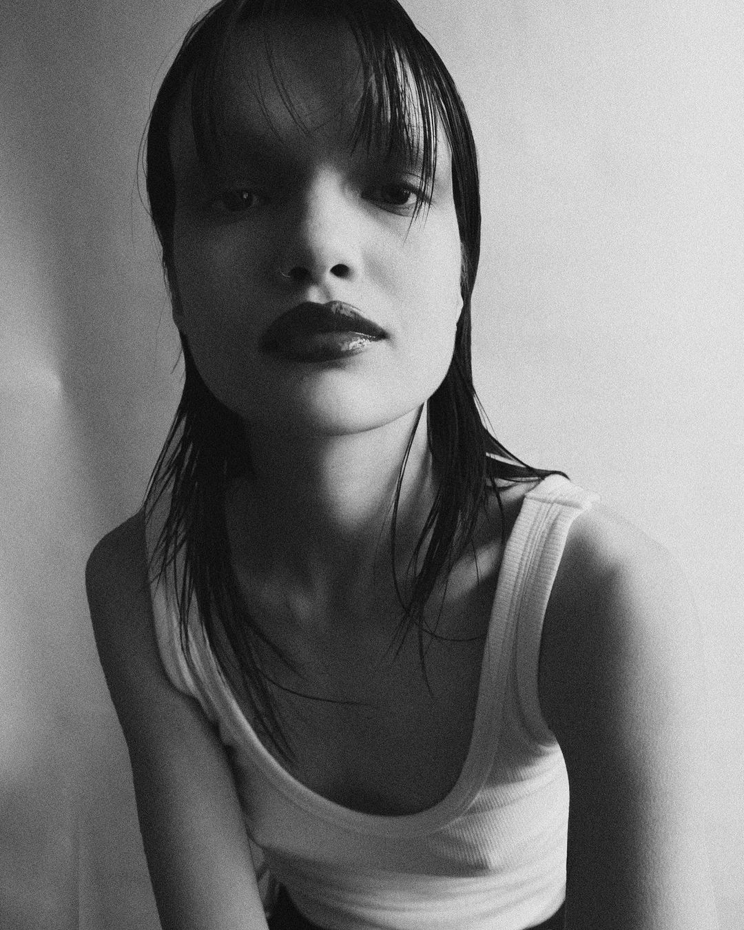 Monochrome portrait of a woman with wet dark hair and glossy dark lipstick, wearing a white tank top against a light grey background.