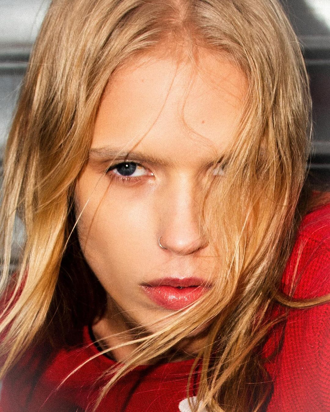 Close-up portrait of a young woman with blonde hair and blue eyes wearing a red sweater, facing forward with a serious expression.