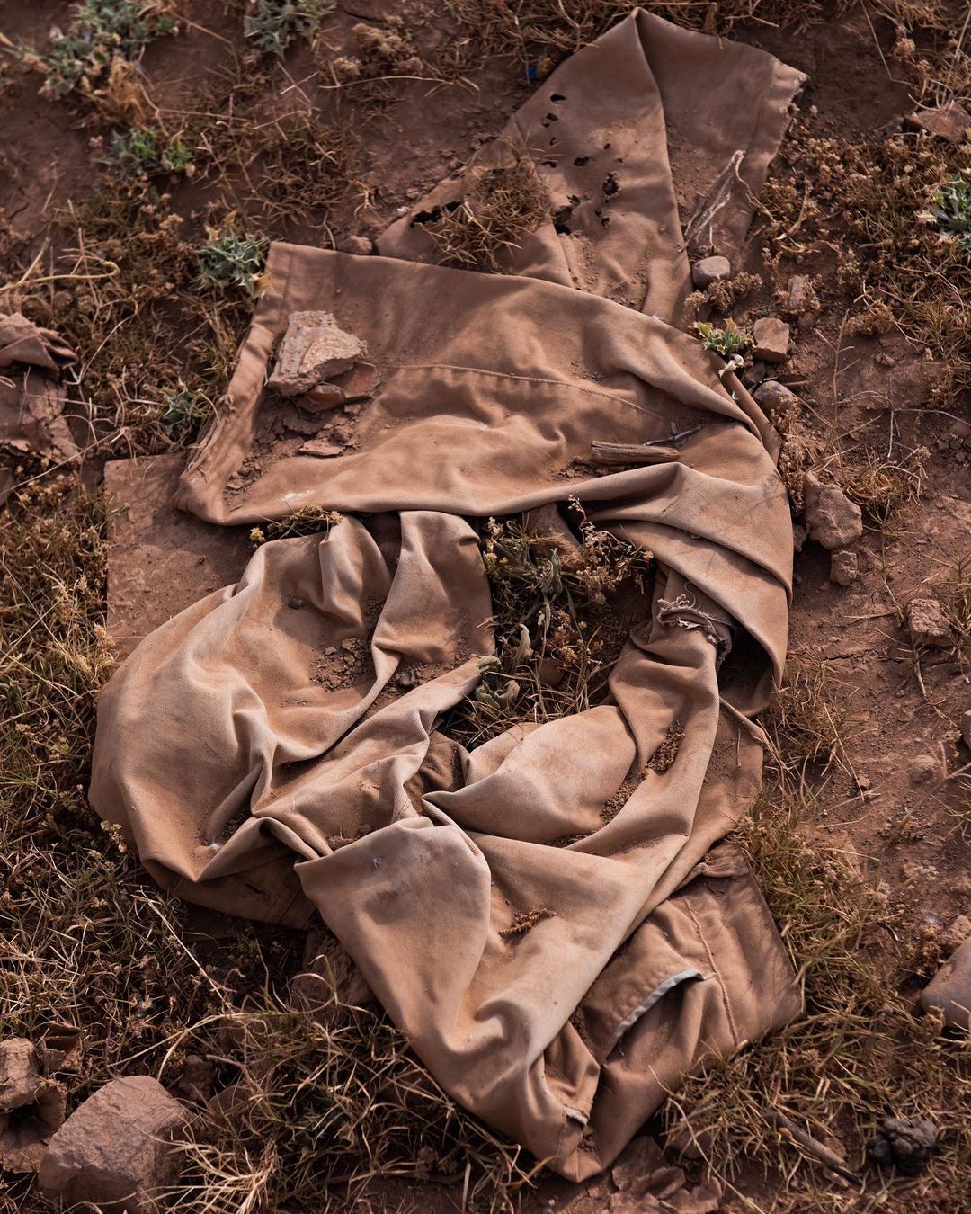 Close-up of a crumpled khaki-colored garment lying on reddish-brown soil, with scattered dry grasses and small green plants visible around it.