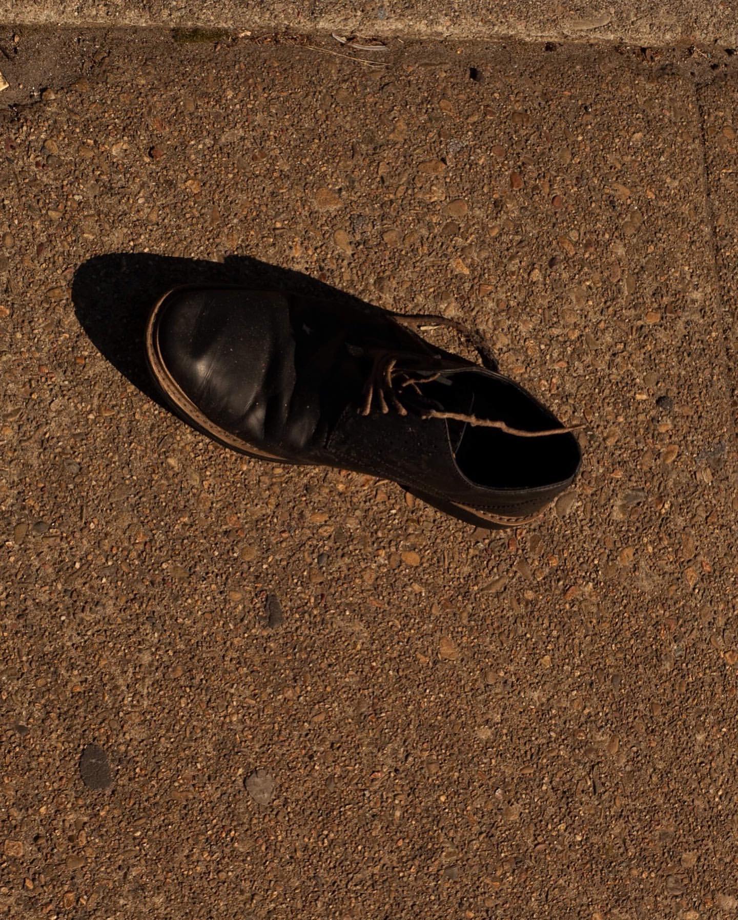 Black leather shoe sits on a concrete sidewalk in the sunlight, with its shadow next to it, creating a striking contrast of light and dark.