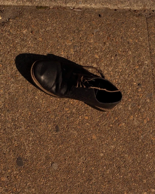 Black leather shoe sits on a concrete sidewalk in the sunlight, with its shadow next to it, creating a striking contrast of light and dark.