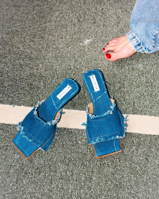 Elevated view of MIISTA denim heeled sandals with frayed edges on a grey textured surface, with a glimpse of a painted toenail.