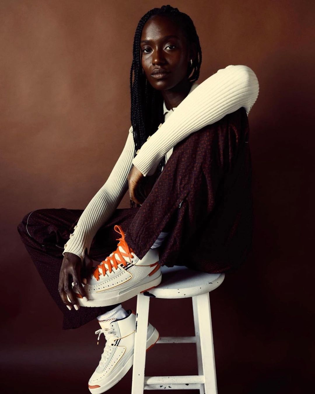 A dark-skinned woman with braids models off-white high-top sneakers with orange laces while sitting on a white stool against a brown backdrop.