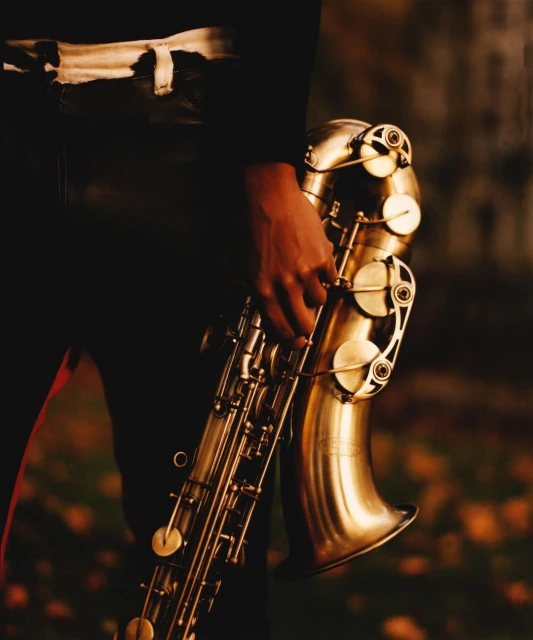 Close up of a person holding a brass saxophone in a moody outdoor setting with blurred background of foliage and muted tones.