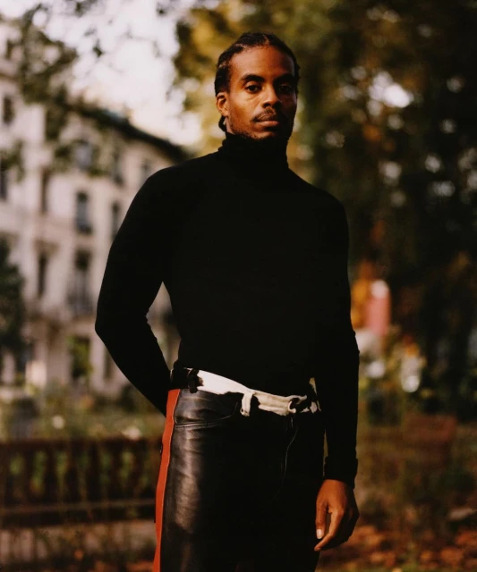 Adult male in a black turtleneck and leather pants poses outdoors in a park-like setting with blurred trees and a building in the background.
