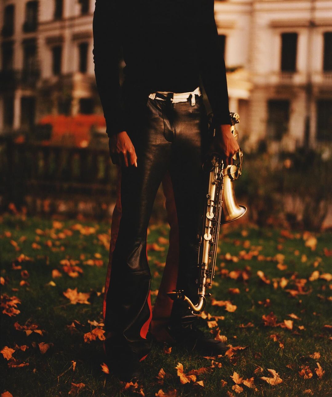 Person in leather pants holding a saxophone on a lawn with autumn leaves near a building, creating a moody, musical ambiance.
