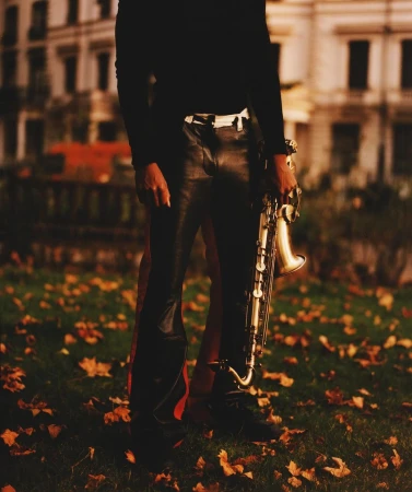 Person in leather pants holding a saxophone on a lawn with autumn leaves near a building, creating a moody, musical ambiance.