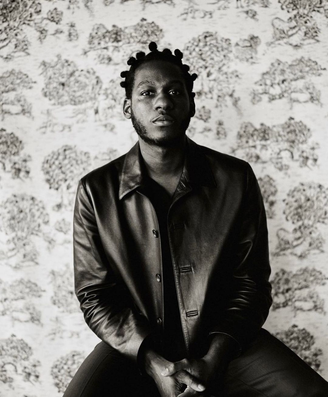 Monochrome portrait of an African American man wearing a leather jacket in front of a decorative patterned wall, looking at the camera.
