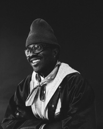 Portrait of a Black man wearing a beanie, glasses, scarf, and jacket against a dark background, photographed by Lawrence Agyei