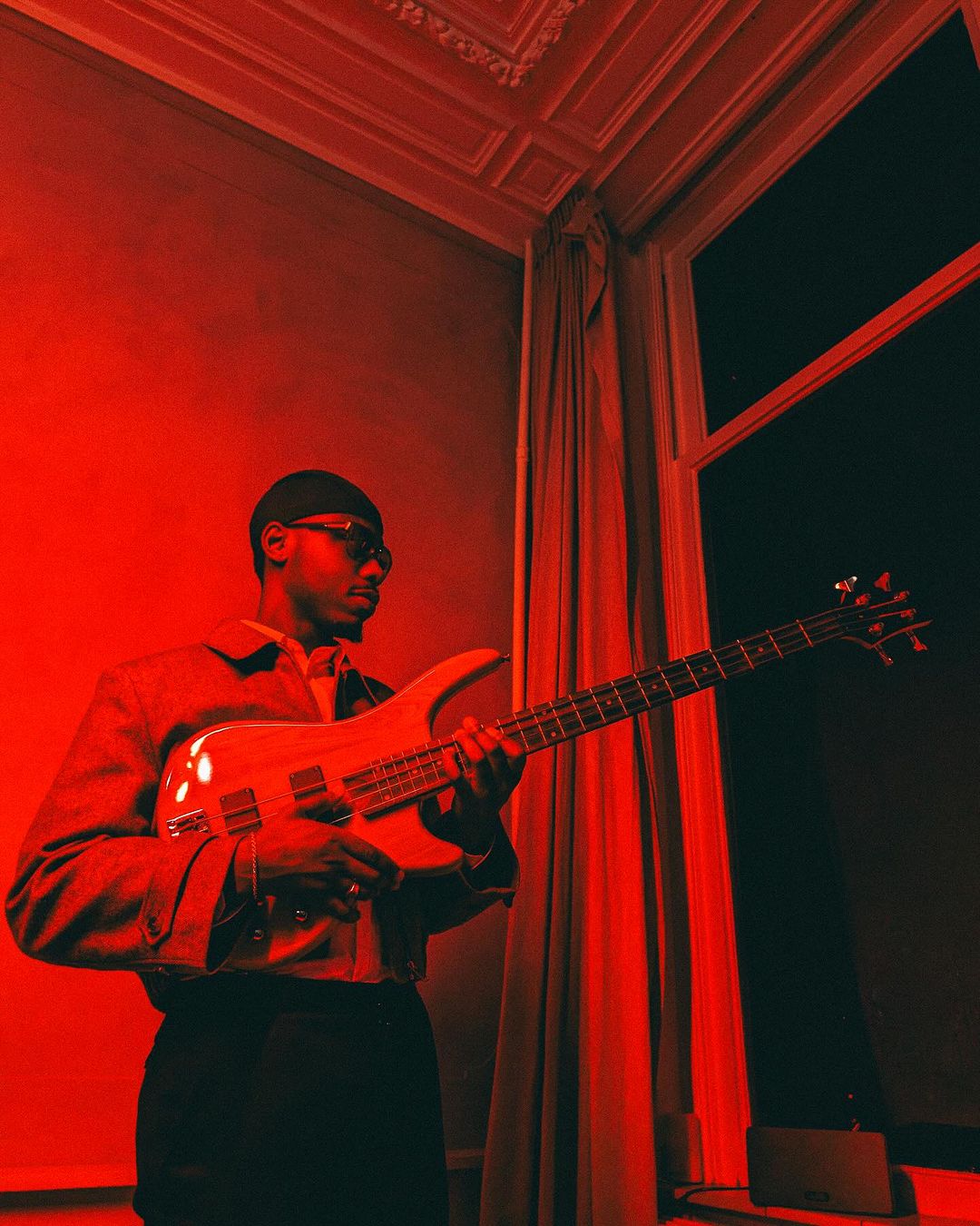 A man plays a bass guitar indoors with red lighting, wearing a jacket, black pants, and a bandana, next to a window with white frame.
