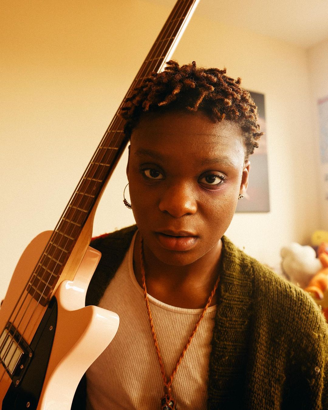 Portrait of a black female musician with dreadlocks, wearing a green cardigan and holding a light pink electric bass guitar indoors.
