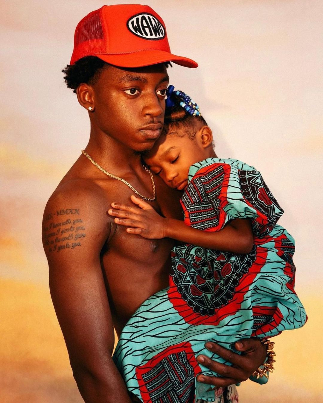 Young man in a red baseball cap cradles a sleeping toddler wearing an Ankara dress against a warm-toned mottled background in a portrait.