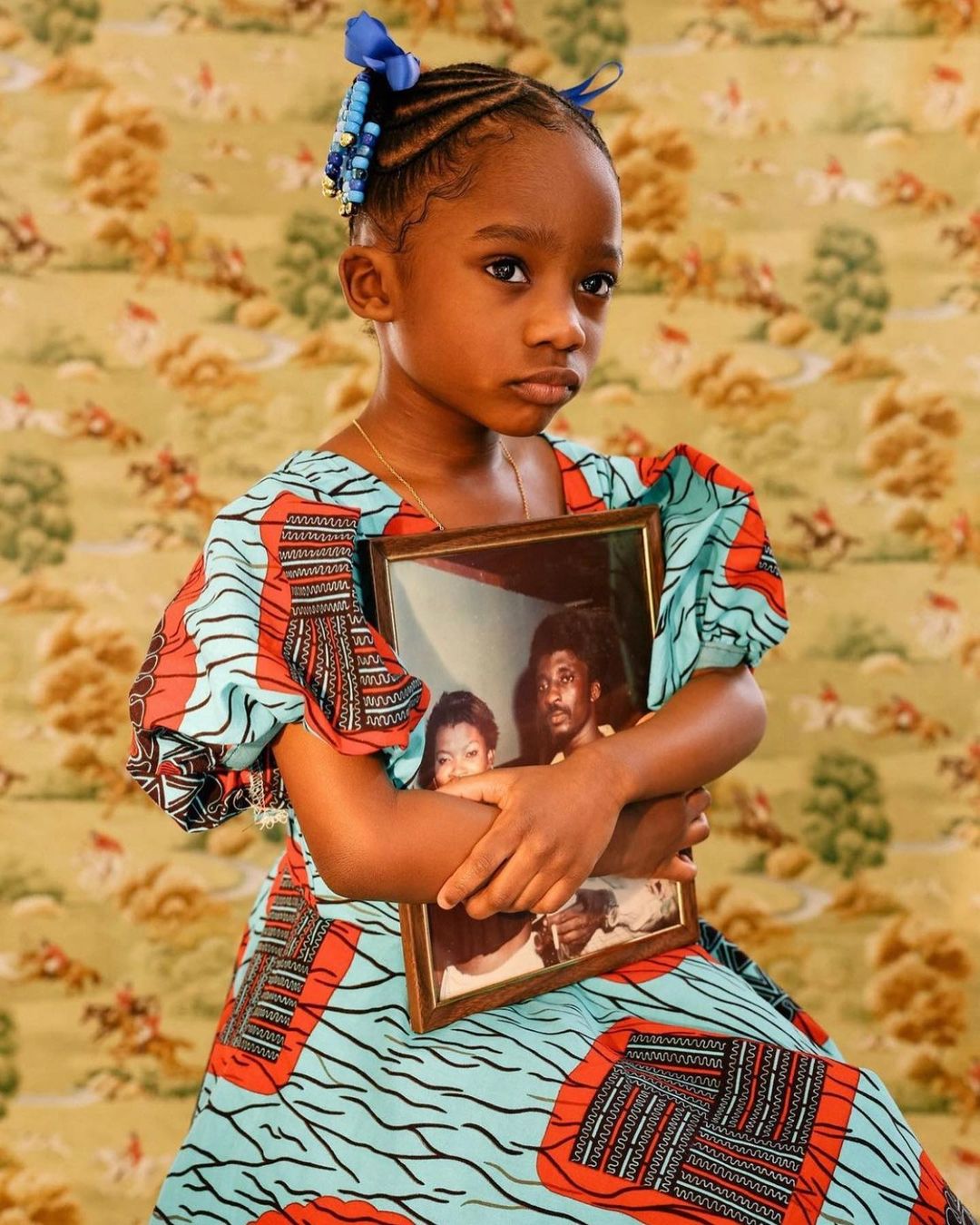 Young girl holds framed photo with sad expression; patterned dress against decorative wallpaper, close-up shot, indoor setting.