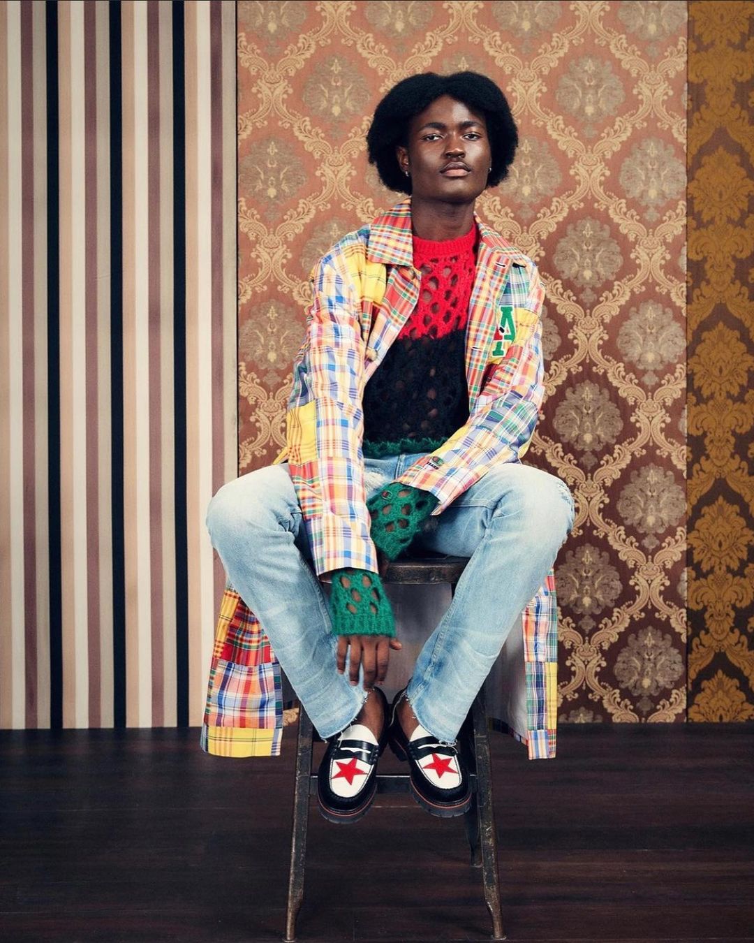Stylish Black man in colorful patchwork coat sits on stool against patterned wallpaper, wearing jeans and loafers.