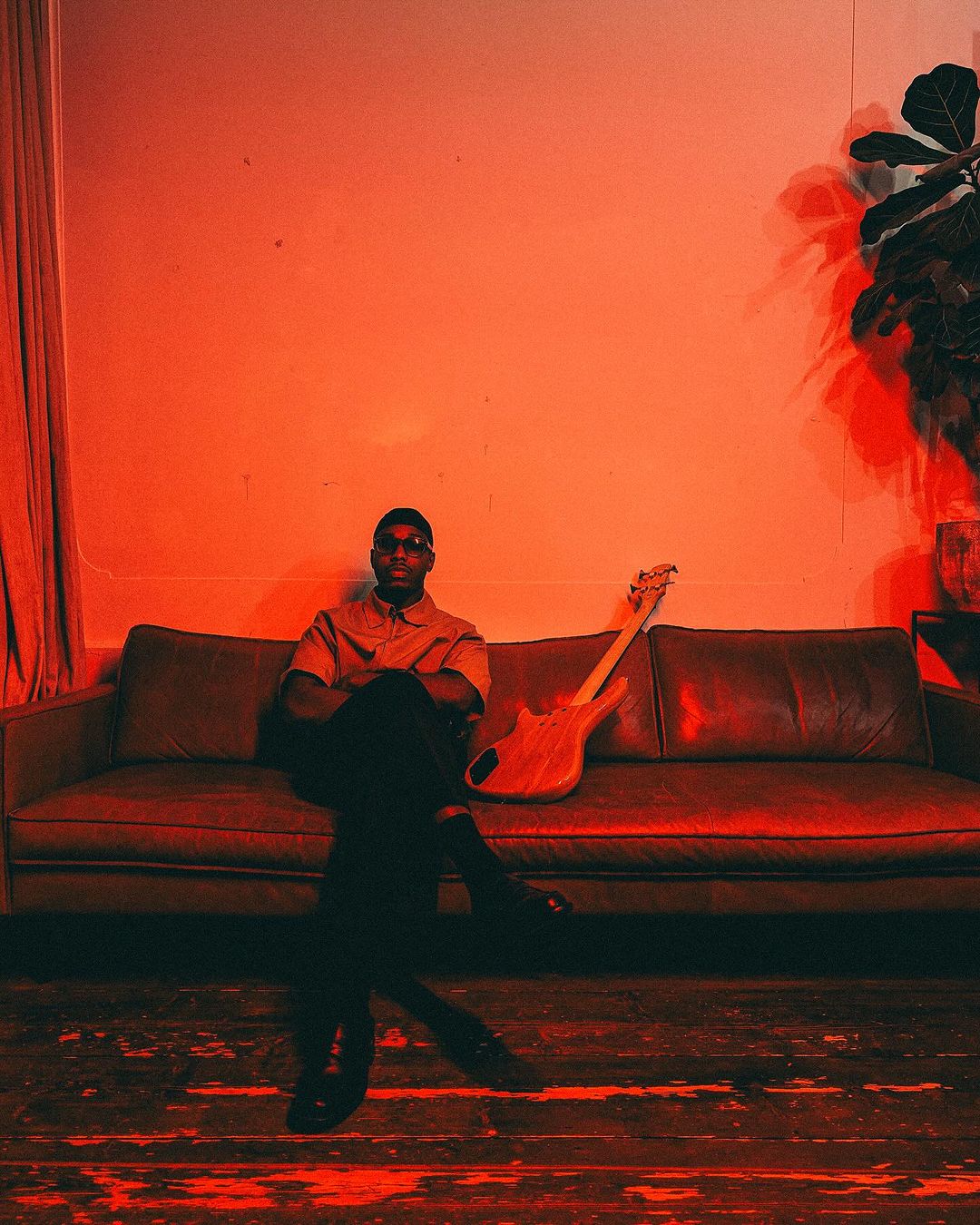An African-American man sitting with crossed legs on a couch with a guitar, wearing sunglasses and a button-down shirt under red lighting.