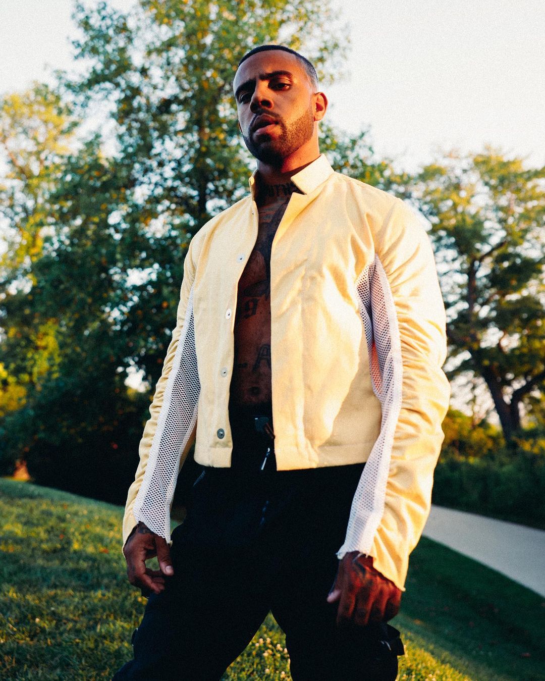 Adult male poses in front of greenery, wearing a yellow jacket and black pants, showcasing fashion and outdoor style in natural light.
