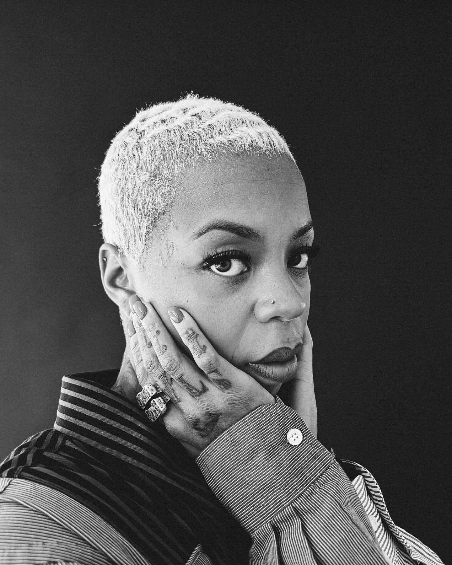 Black woman with short dyed blonde hair and tattoos on her fingers posing against a dark background in a portrait photograph by Lawrence Agyei