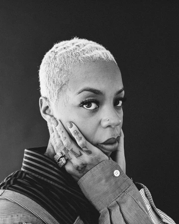 Black woman with short dyed blonde hair and tattoos on her fingers posing against a dark background in a portrait photograph by Lawrence Agyei