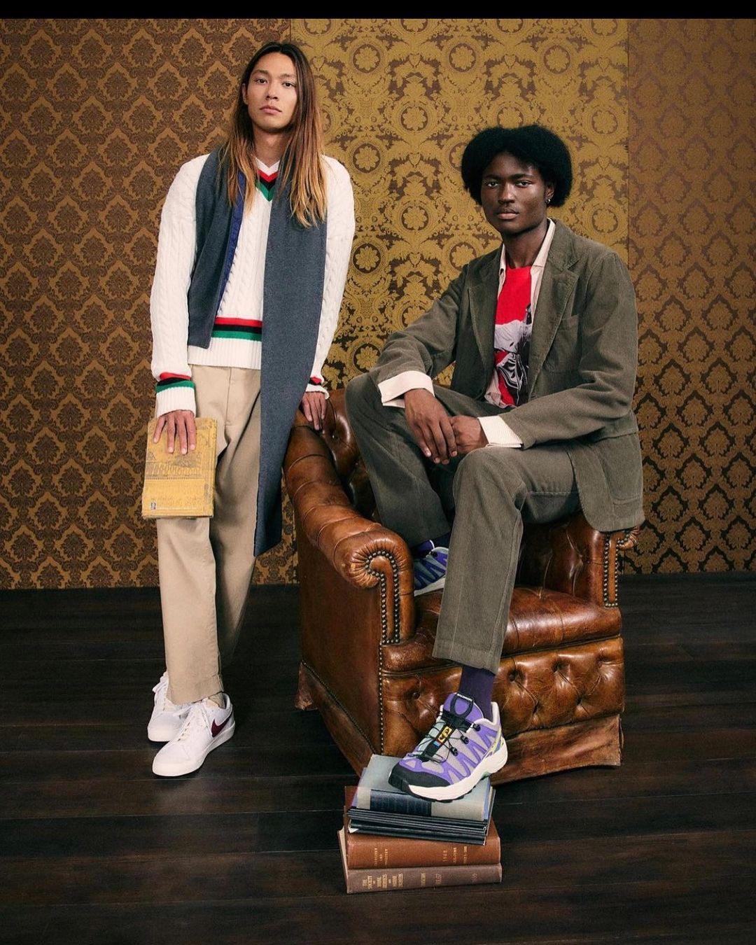 Two male fashion models pose in a vintage-styled studio setting with books and a brown leather armchair, wearing coordinated outfits.