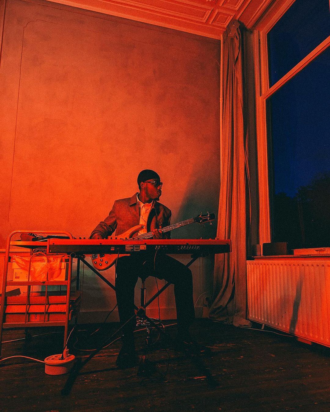 Adult male musician plays keyboard and guitar in a room with orange walls, window, and radiator, illuminated by orange light.