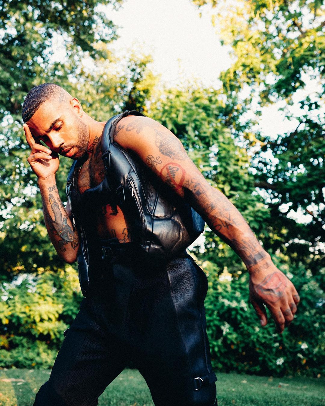 Adult male with tattoos in black vest and pants poses in a natural outdoor setting with green trees and dappled lighting.