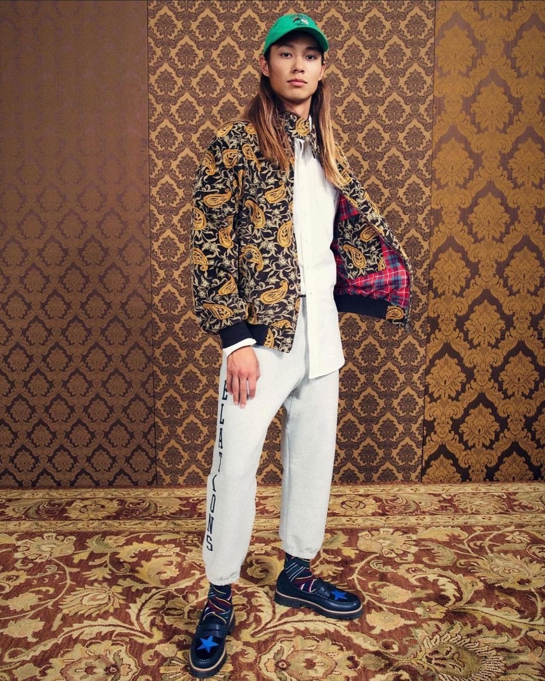 Stylish individual poses in patterned jacket, white shirt, sweatpants, and blue shoes against an ornate patterned backdrop and carpet.